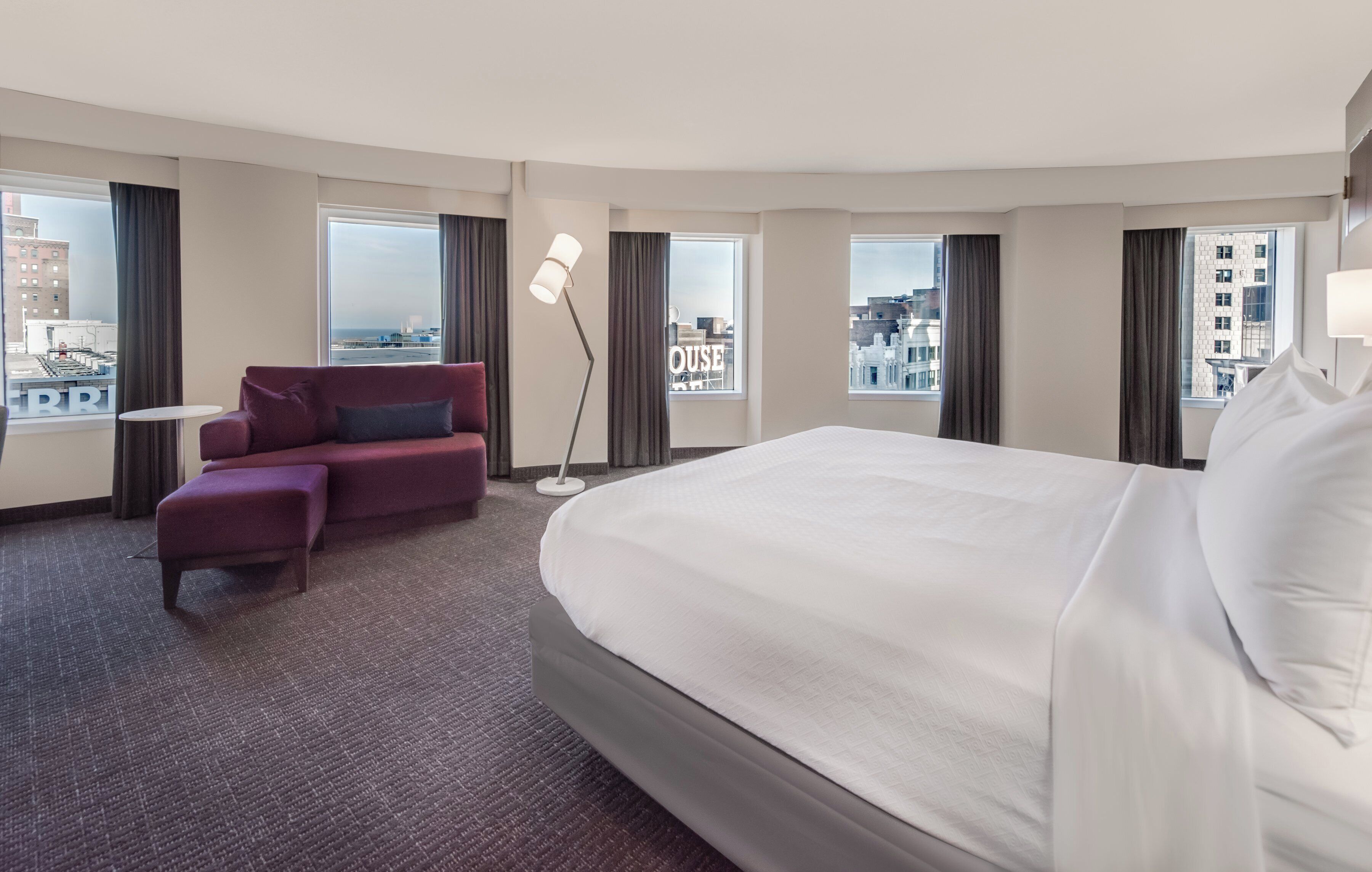 A hotel room with a large bed, a sofa, a small table, a floor lamp, and large windows with city views.