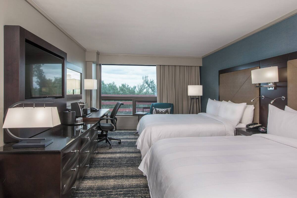 A hotel room with two beds, a desk with a chair, a TV, and a large window with curtains.