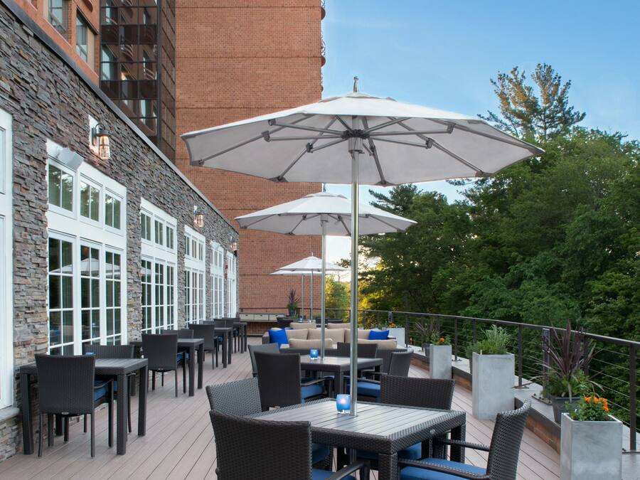 Een terras buiten met tafels en stoelen, parasols, en een bakstenen gebouw op de achtergrond.