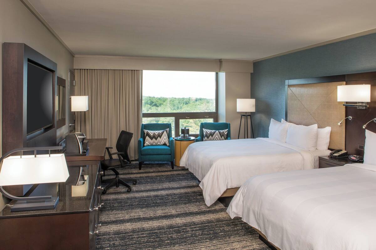 A hotel room with two beds, a desk with a computer, a TV, and a window with a view of greenery.