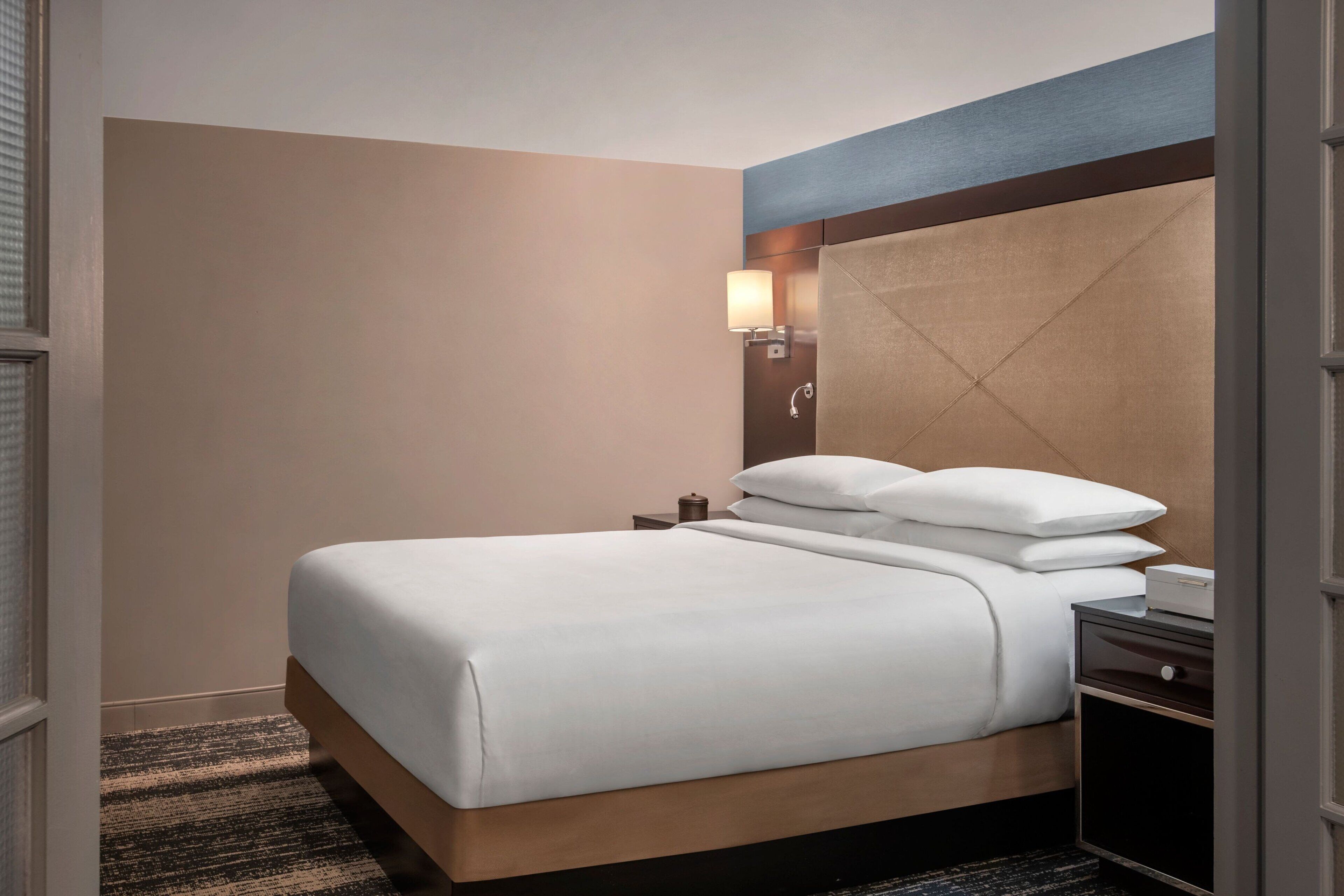 A neatly made bed with white linens, a wooden headboard, a nightstand with a lamp, and a small drawer.
