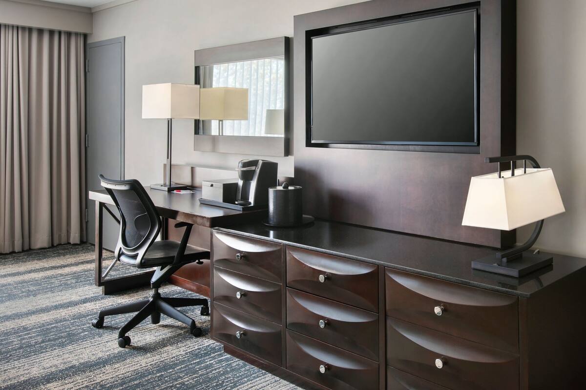 A hotel room with a dark wood dresser, a desk with a chair, a flat-screen TV, and two lamps.