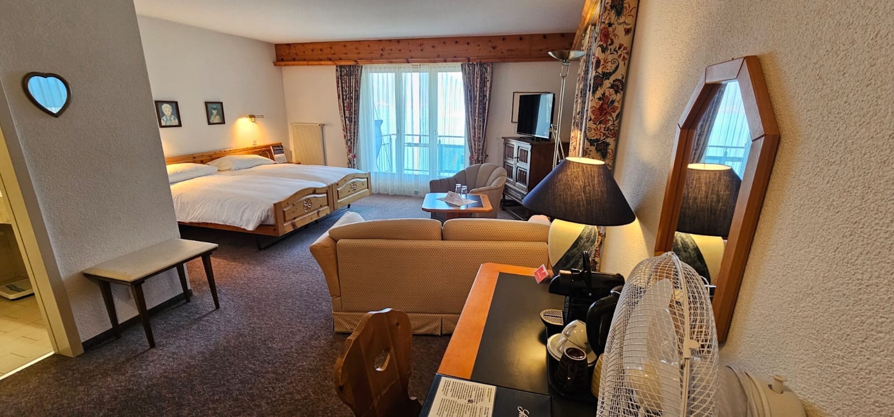 A hotel room with a bed, a sofa, a desk with a lamp, a chair, a small table, and a television.