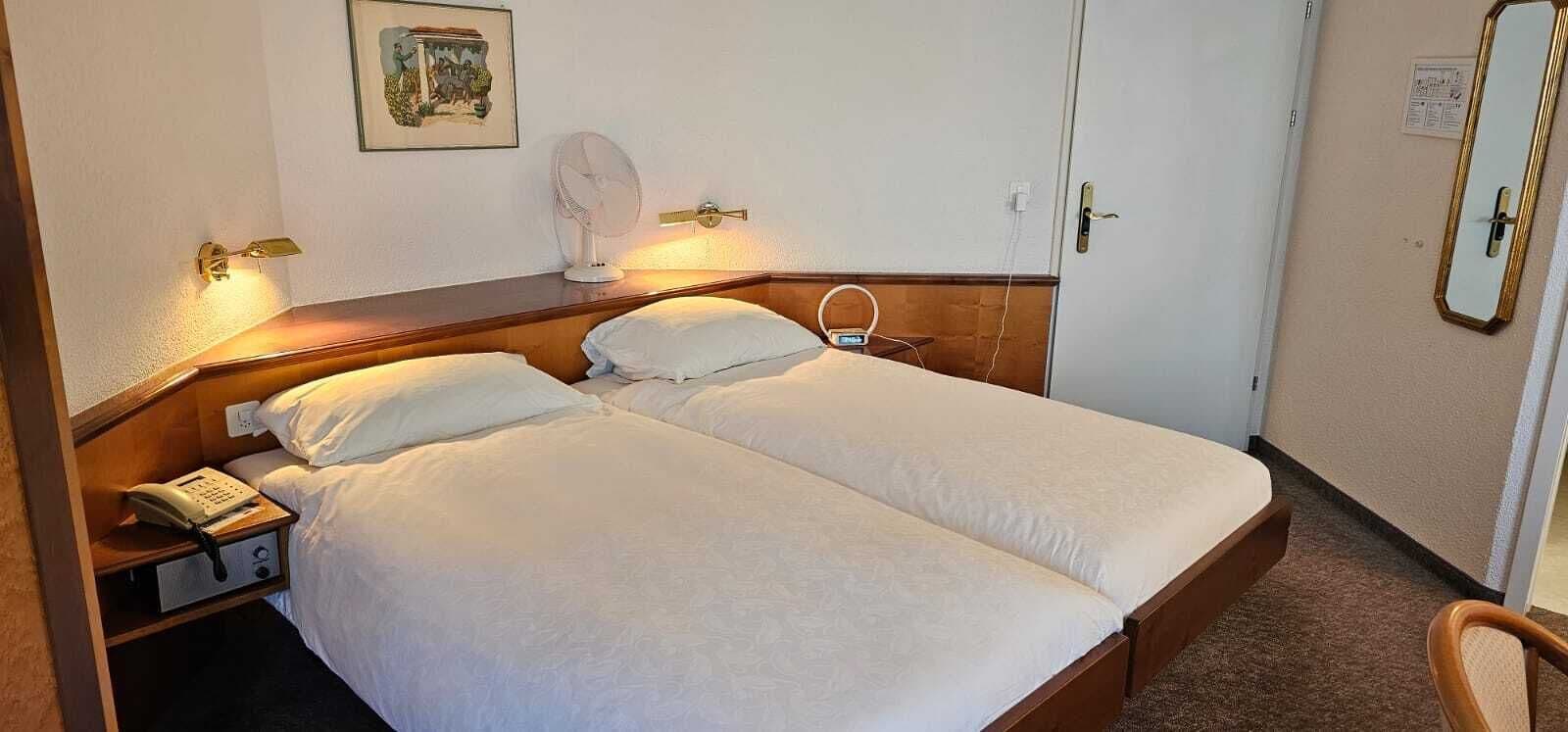 Een tweepersoonsbed met witte lakens, een nachtkastje met een telefoon, een ventilator aan de muur en een spiegel.