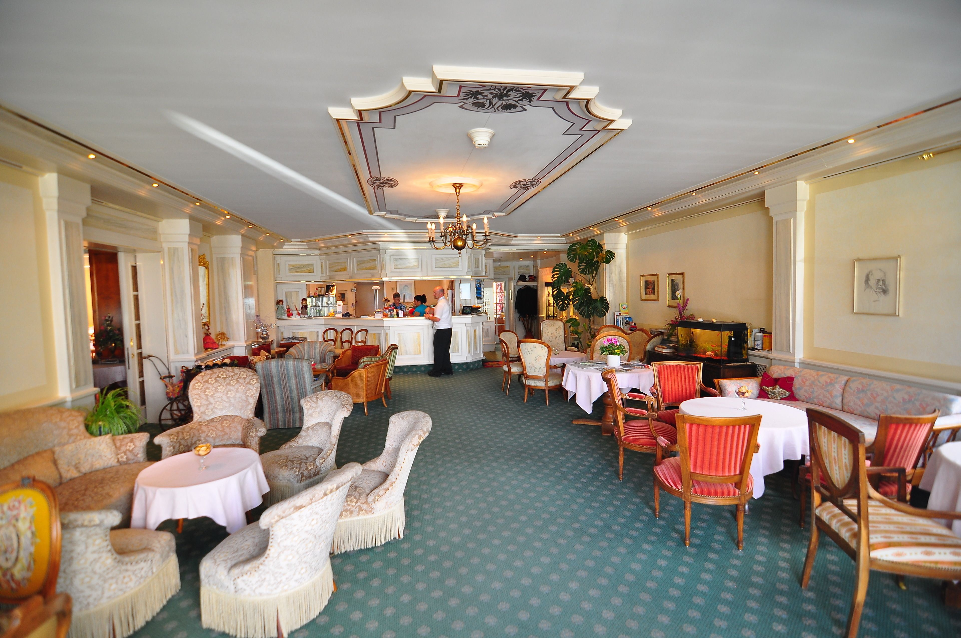 Un amplio vestíbulo de hotel con una zona de bar central, varios arreglos de asientos que incluyen mesas redondas y sillas, y una alfombra estampada.
