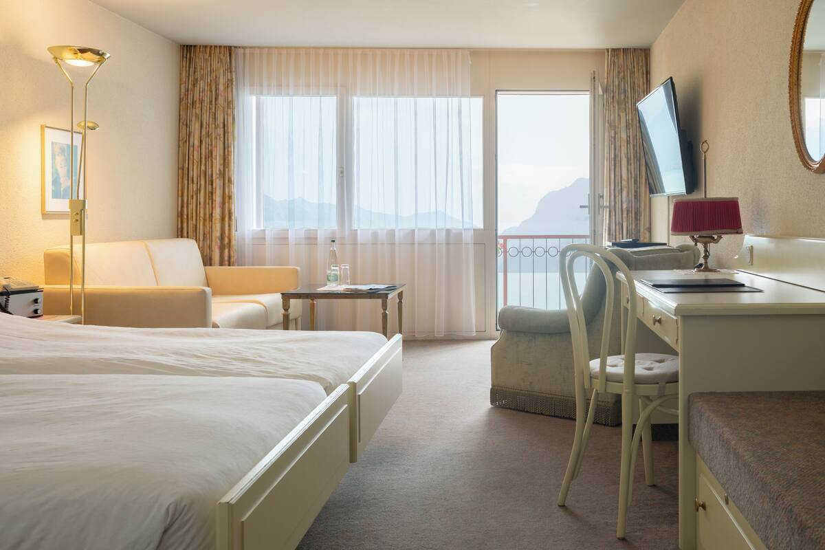 A hotel room with two beds, a desk, a chair, a TV, and a balcony with a view.