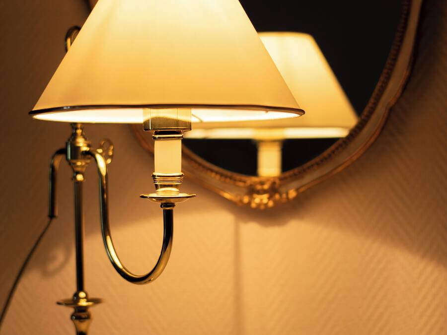 A table lamp with a white shade and a gold base, positioned in front of a mirror.