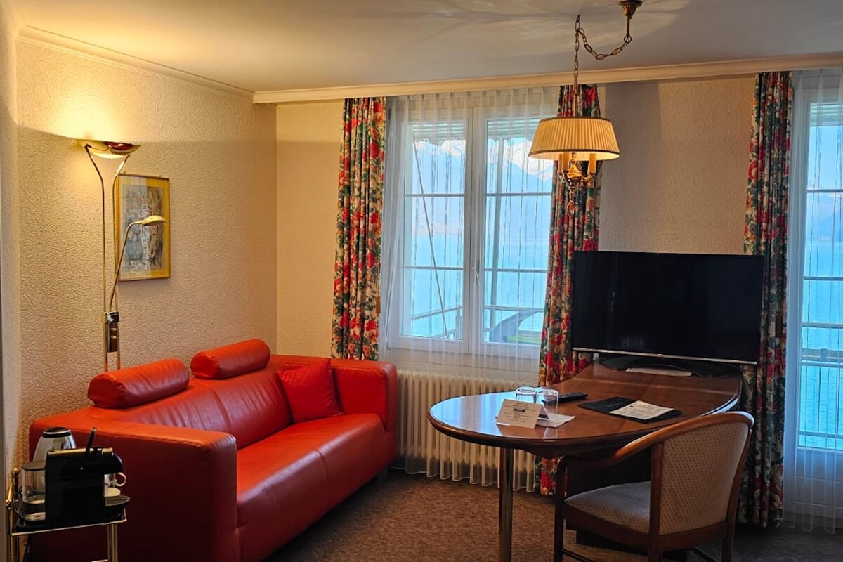 A hotel room with a red leather sofa, a television, a small table with a chair, and a window with curtains.
