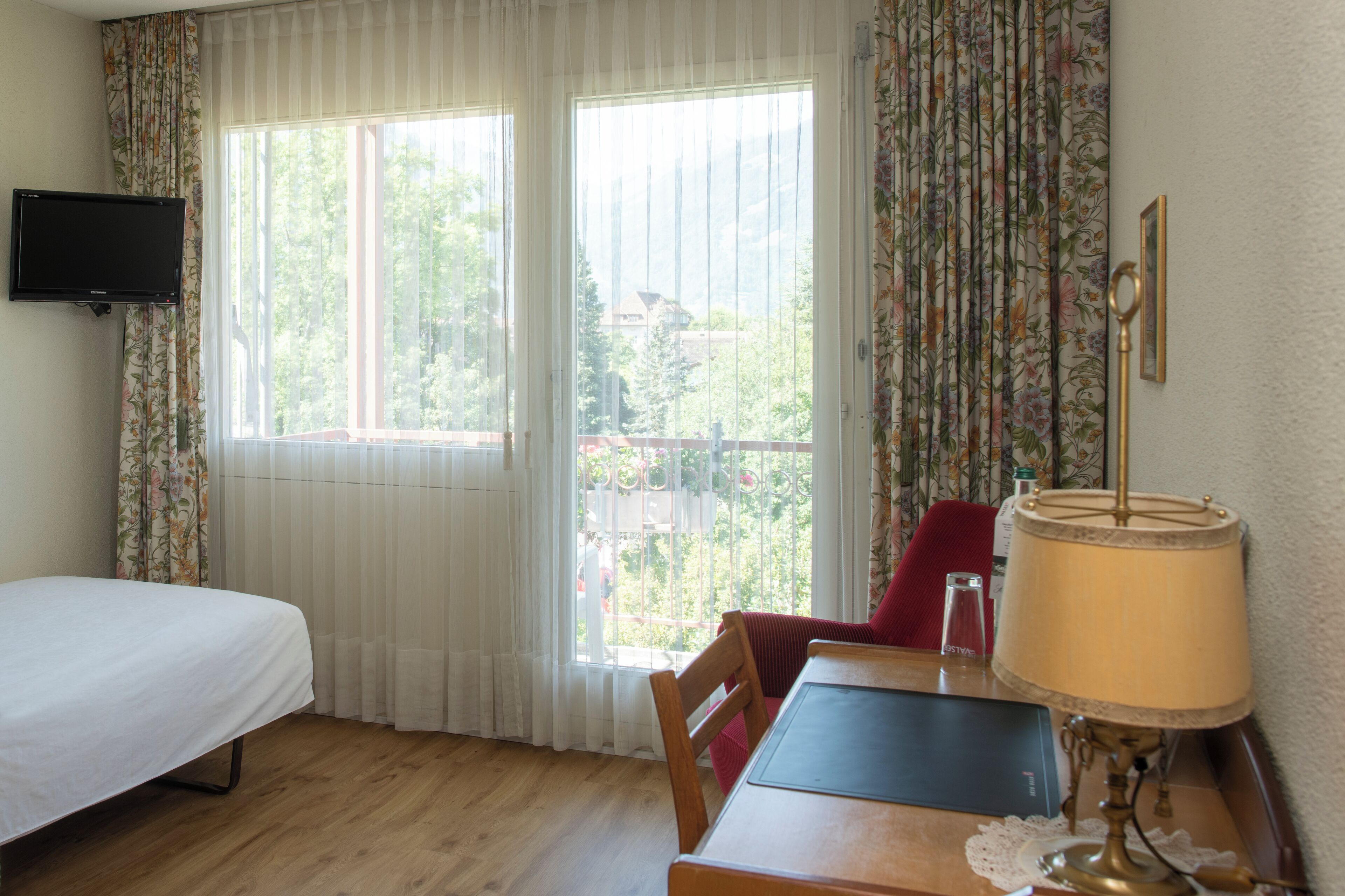 A room with a bed, a table with a lamp, a chair, and a television on the wall.