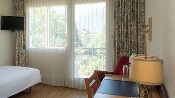 A room with a bed, a table with a lamp, a chair, and a television on the wall.