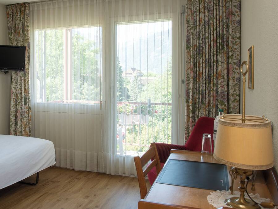 A room with a bed, a table with a lamp, a chair, and a television on the wall.