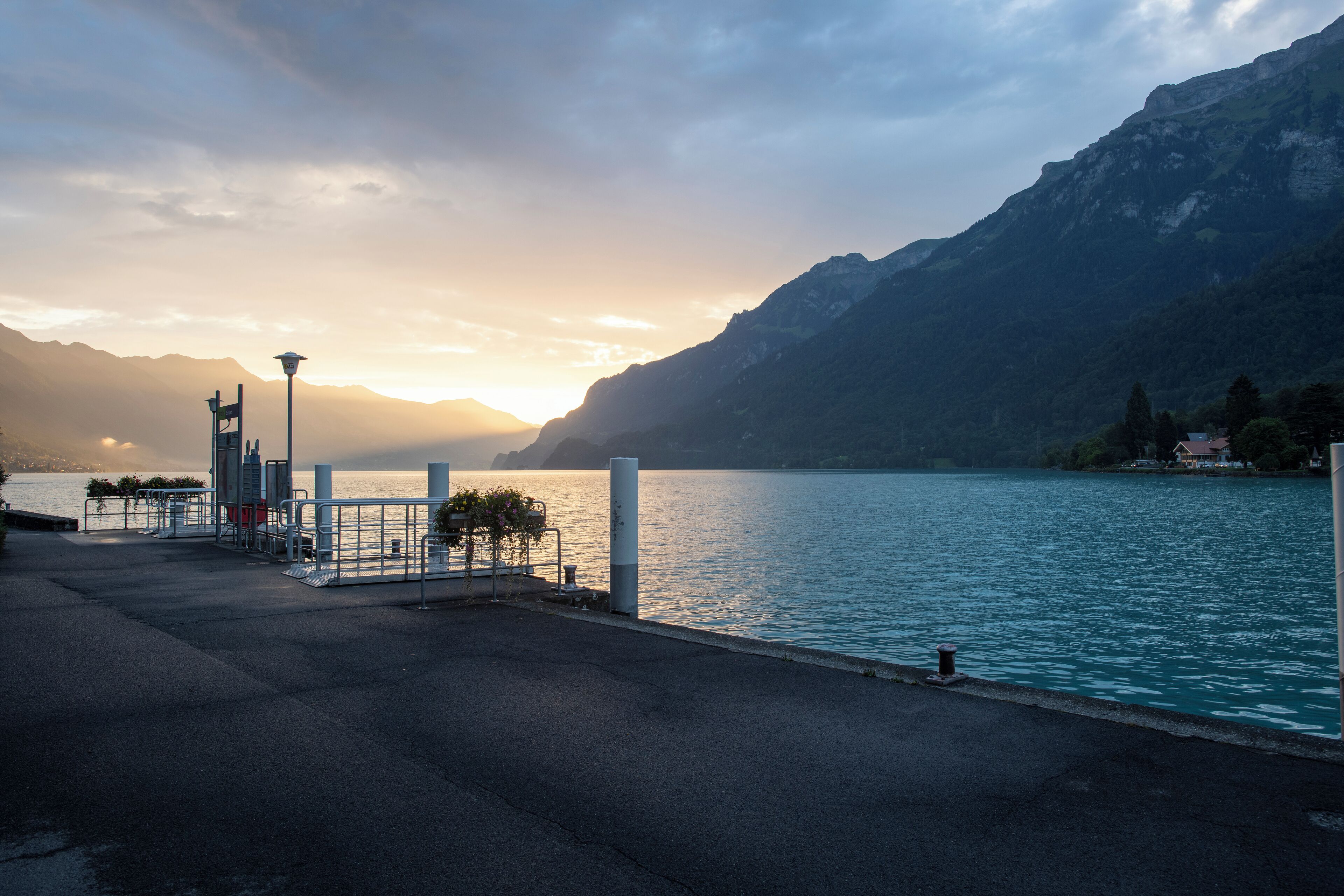 Eine Uferpromenade mit einer Laterne, Bänken und Blick auf einen See und Berge.