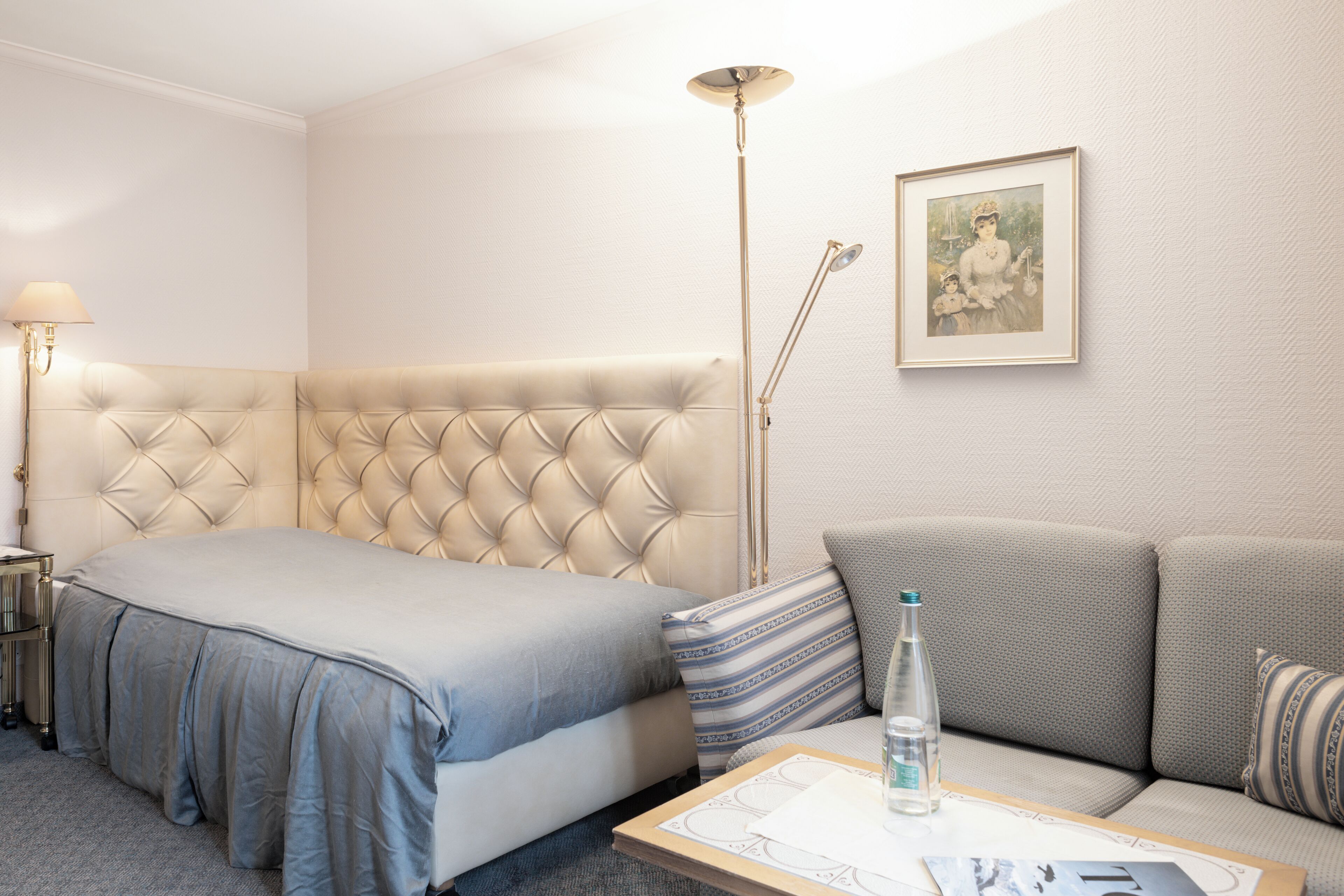 Une chambre d’hôtel avec un lit doté d’une tête de lit capitonnée, un canapé, une lampe sur pied et un tableau encadré au mur.