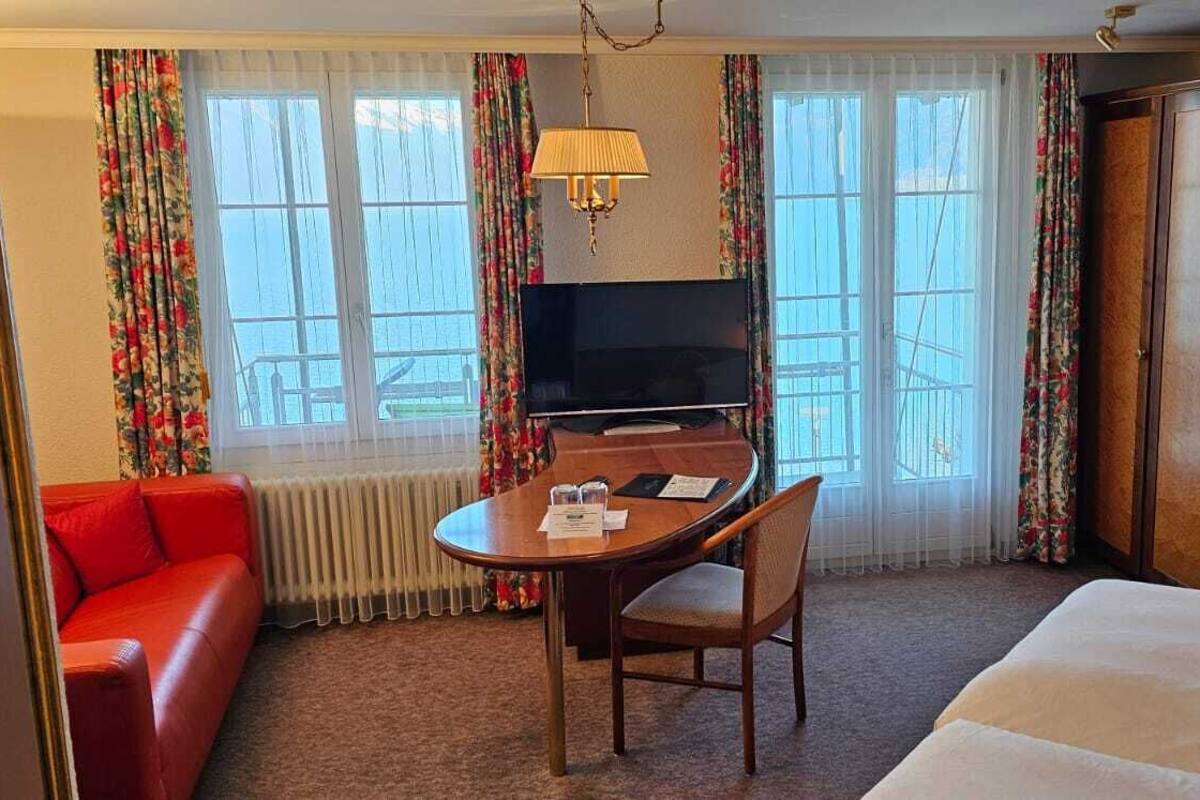 A hotel room with a bed, a red sofa, a dining table with chairs, a TV, and a wardrobe.