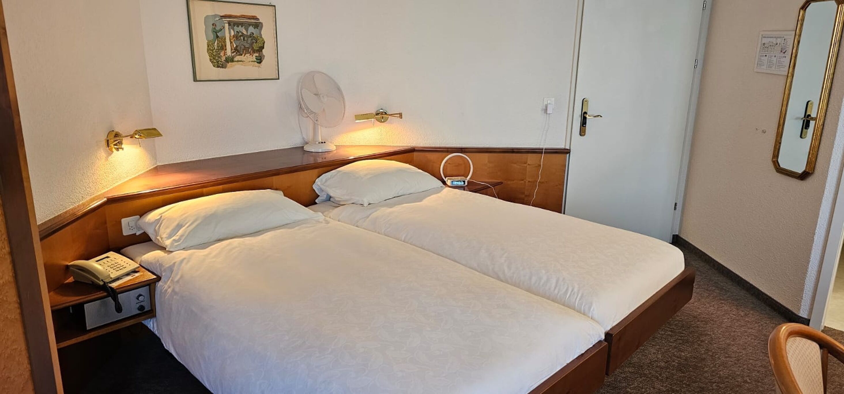 A double bed with white linens, a bedside table with a telephone, a wall-mounted fan, and a mirror.
