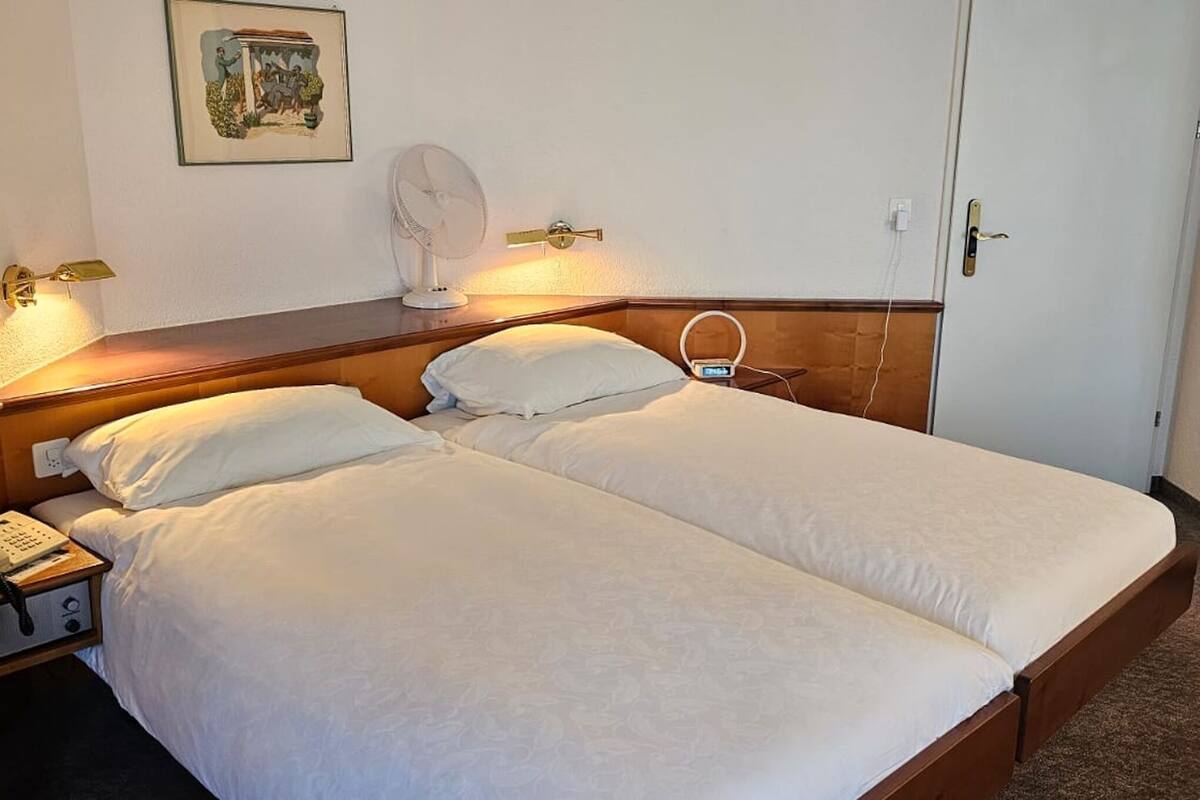 A double bed with white linens, a bedside table with a telephone, a wall-mounted fan, and a mirror.