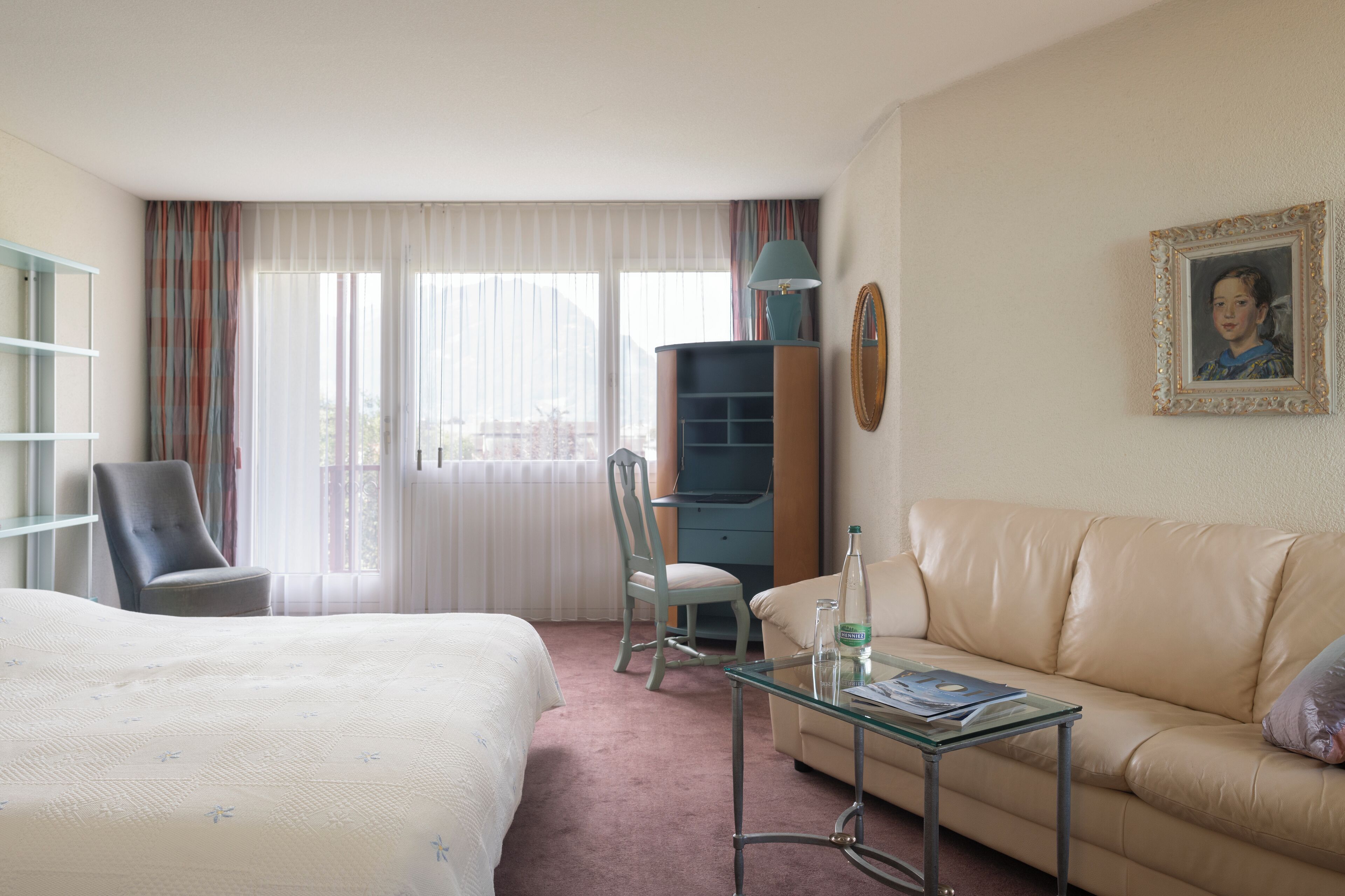 Quarto de hotel com cama, sofá, mesa de centro de vidro, cadeira, escrivaninha e janela com cortinas.