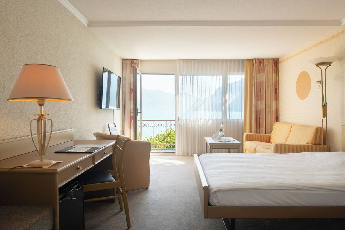 A hotel room with a bed, desk, chair, and a view of the outdoors.