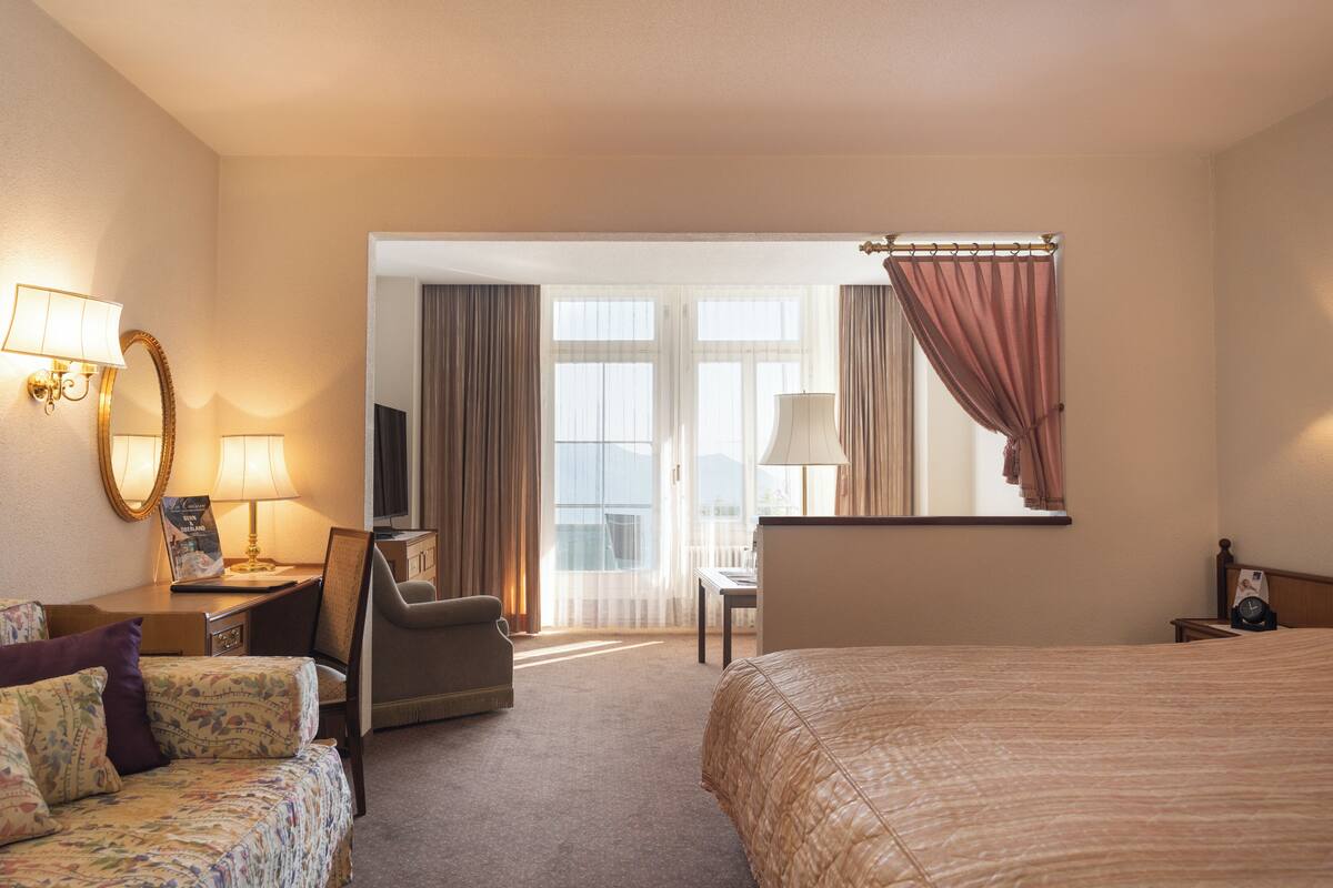 A hotel room with a bed, a desk with a chair, a sofa, a TV, and a window with curtains.