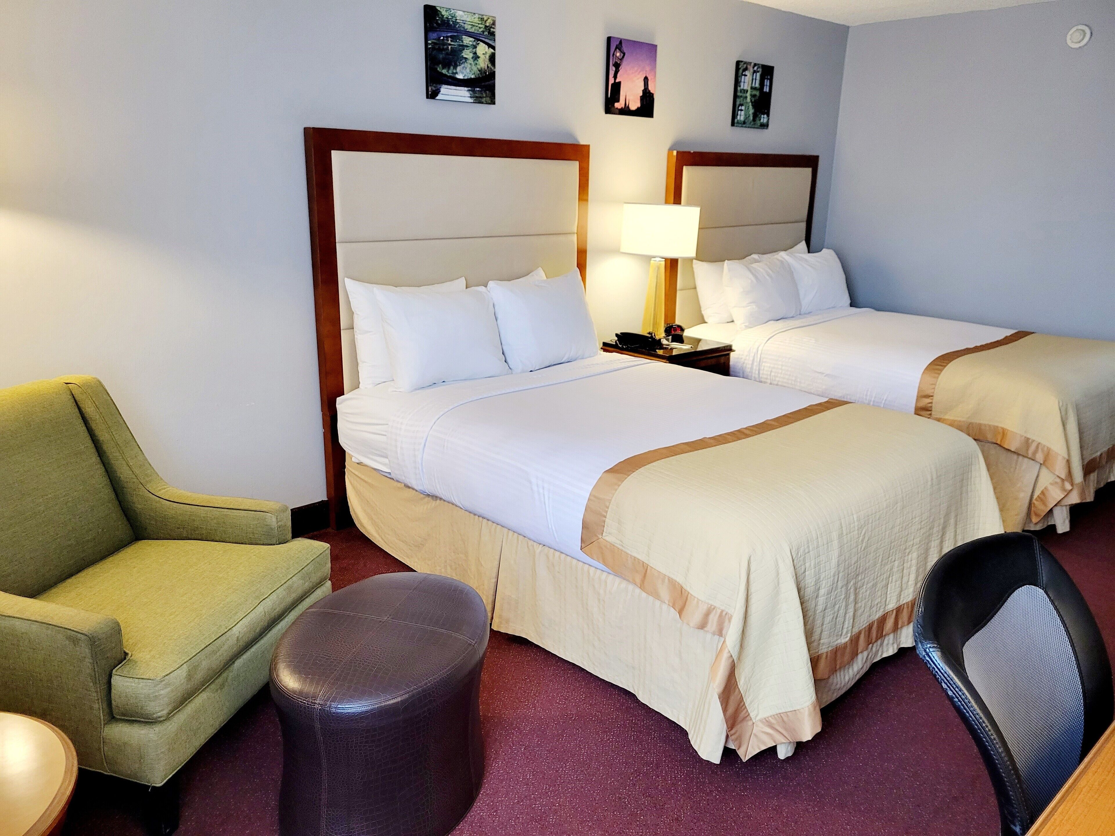 Une chambre d’hôtel avec deux lits, un fauteuil vert, un petit pouf rond et un bureau avec une chaise.