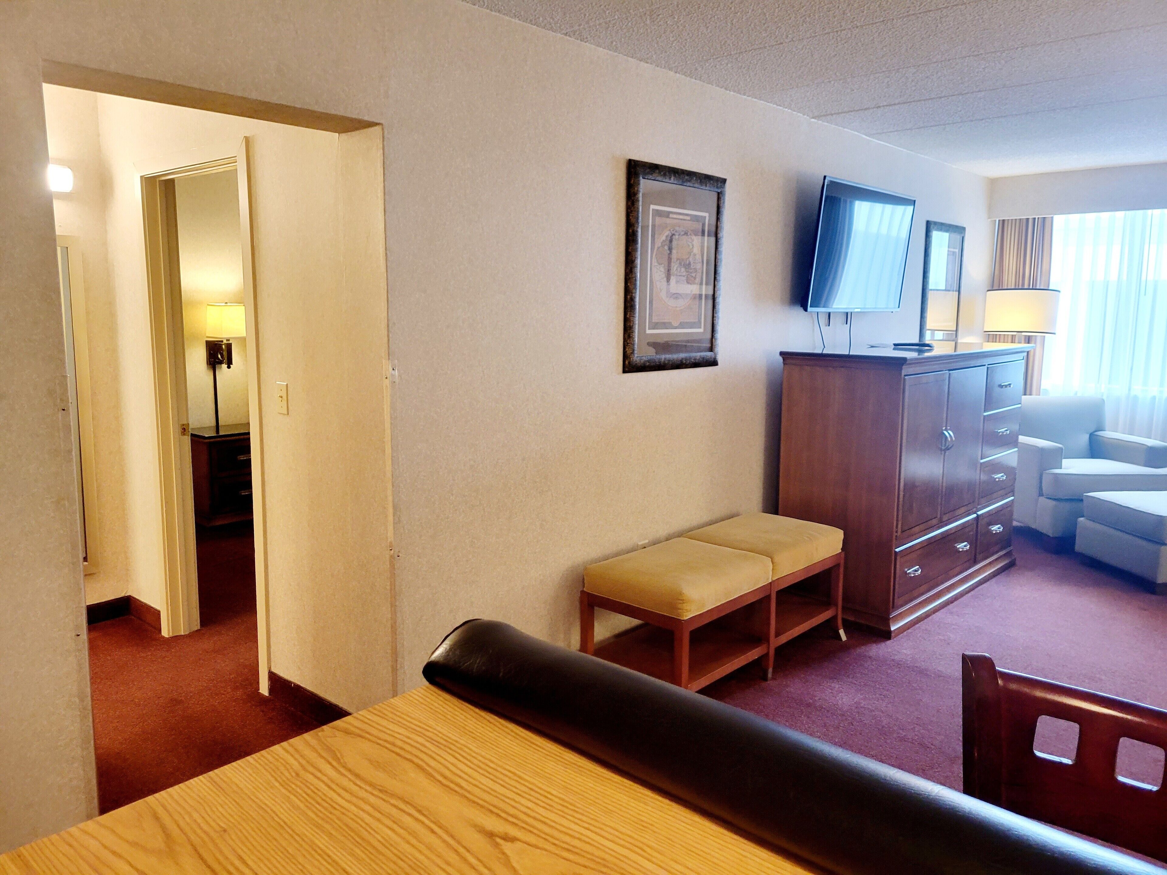 A hotel room with a wooden desk, a bench, a television on a cabinet, and a door leading to another room.