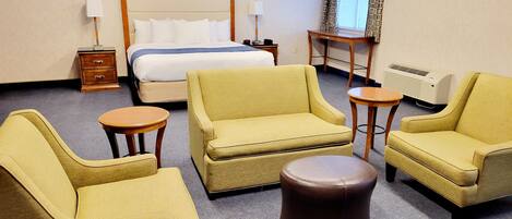 Une chambre d’hôtel comprenant un lit, deux fauteuils, un petit pouf rond, une table de chevet, une table et une fenêtre avec des rideaux.