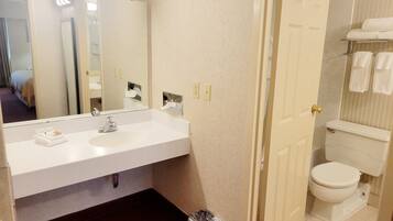 A bathroom with a white sink, mirror, and toilet. There is a trash can and a towel rack with towels.