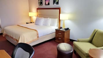 A hotel room with a bed, bedside lamps, a desk, a chair, a sofa, and a small round ottoman.