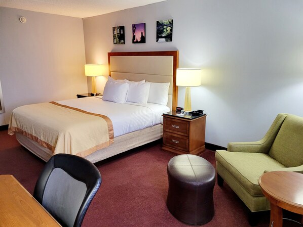 Una habitación de hotel con cama, lámparas de noche, escritorio, silla, sofá y un otoman redondo pequeño.