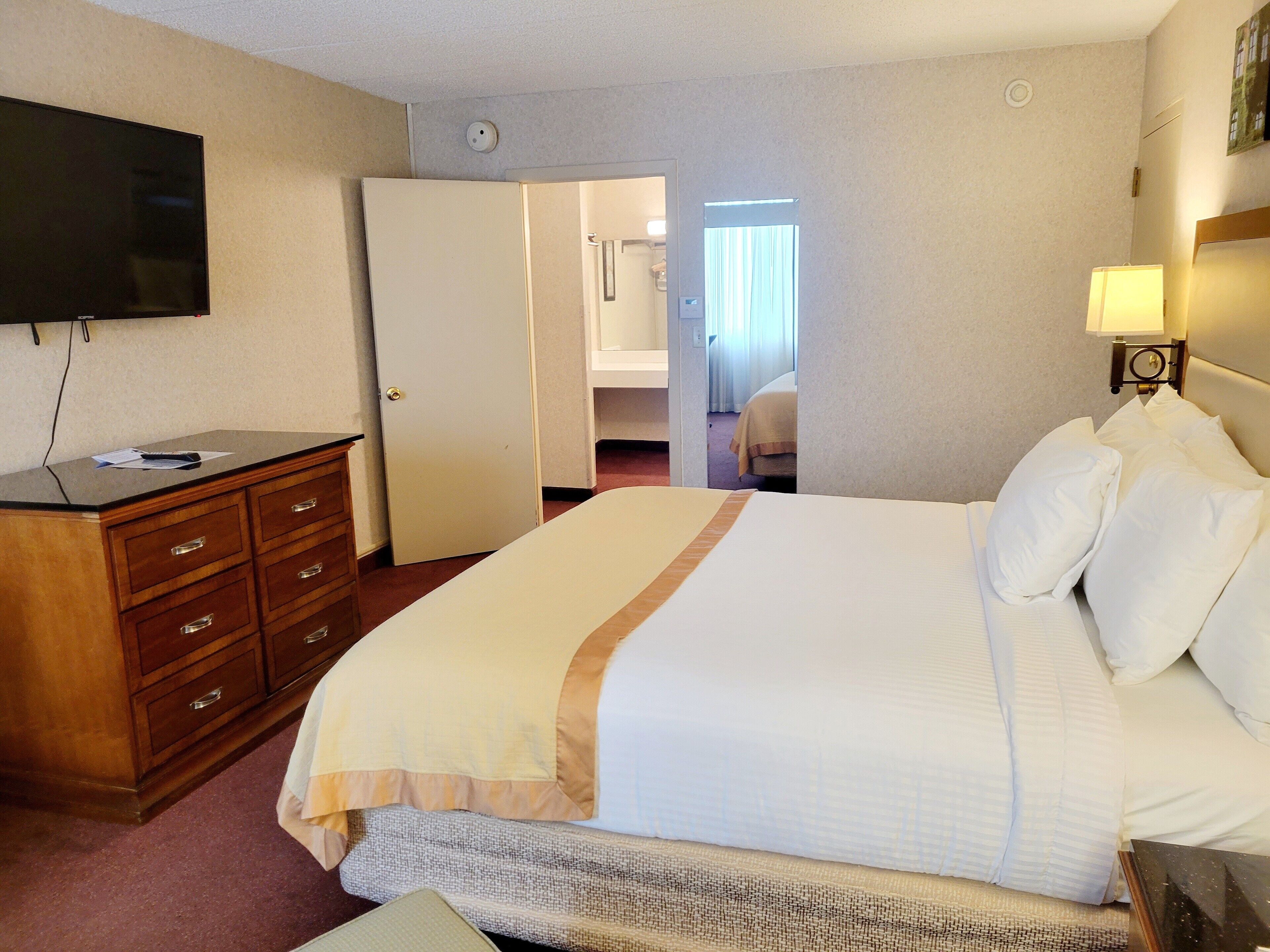 A hotel room with a bed, a dresser, a television, and a door leading to another room.