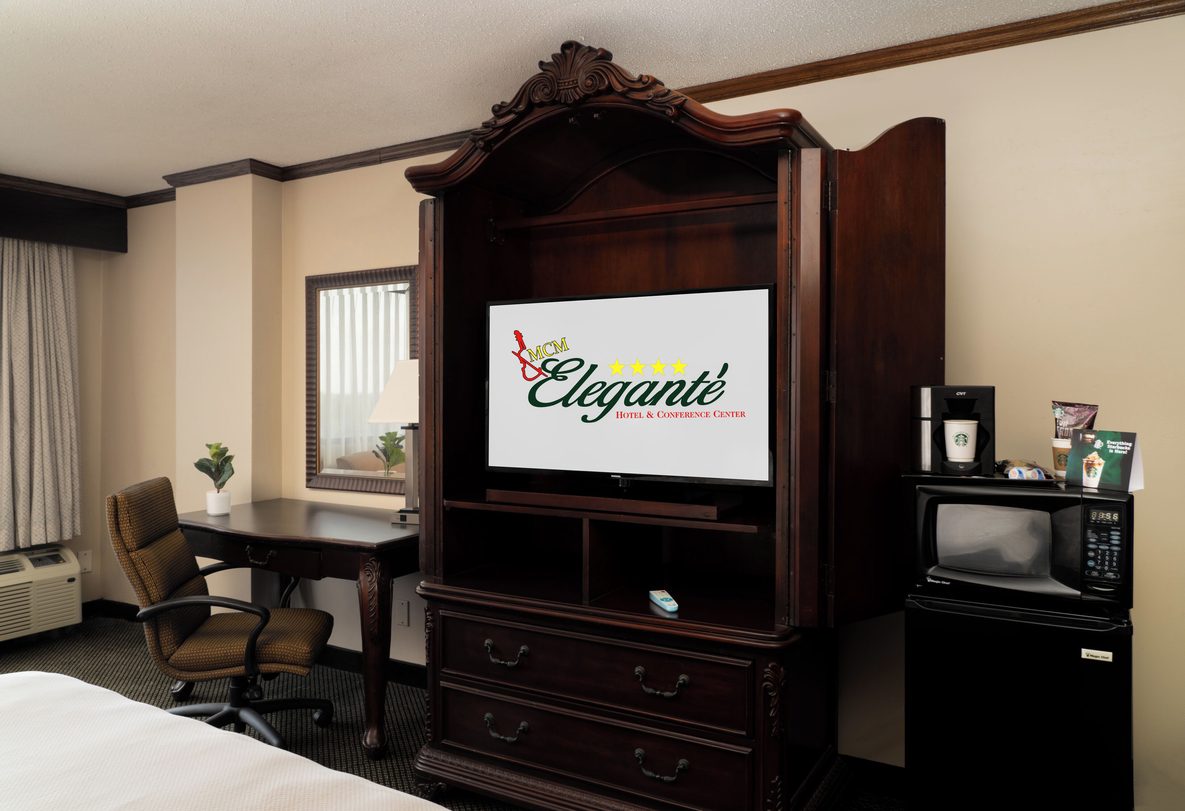 A hotel room with a large flat-screen TV, a wooden entertainment center, a desk with a chair, a microwave, and a coffee maker.