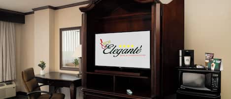 A hotel room with a large flat-screen TV, a wooden entertainment center, a desk with a chair, a microwave, and a coffee maker.
