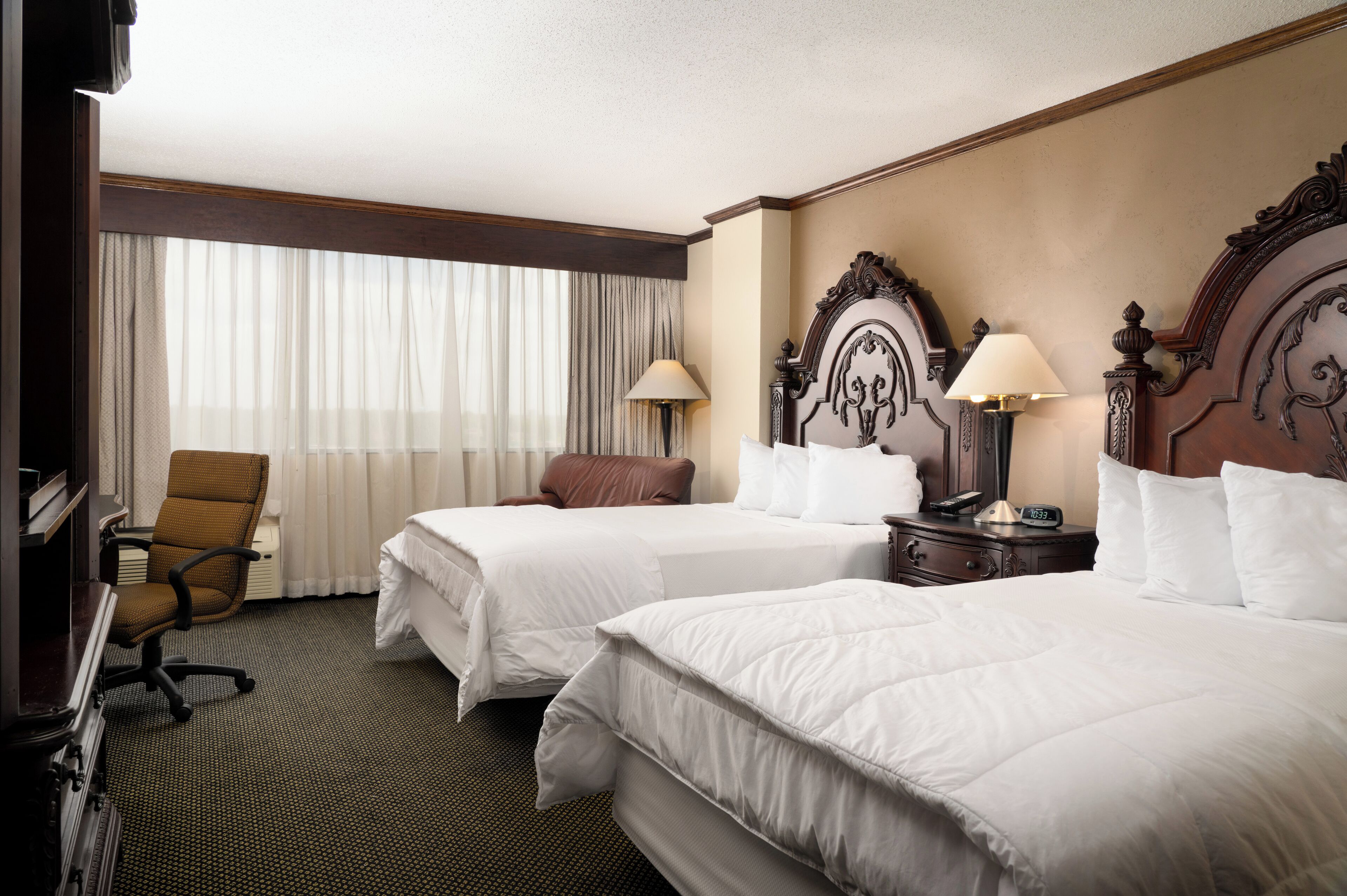 A hotel room with two beds, a desk, a chair, and a large window with curtains.