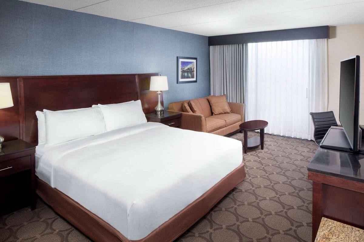 Ein Hotelzimmer mit einem großen Bett, einer Couch, einem Schreibtisch mit Fernseher und einem Fenster mit Vorhängen.