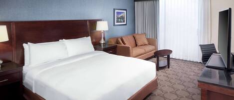 A hotel room with a large bed, a sofa, a desk with a television, and a window with curtains.