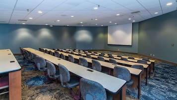 A conference room with rows of tables and chairs, a projector screen, and a carpeted floor.