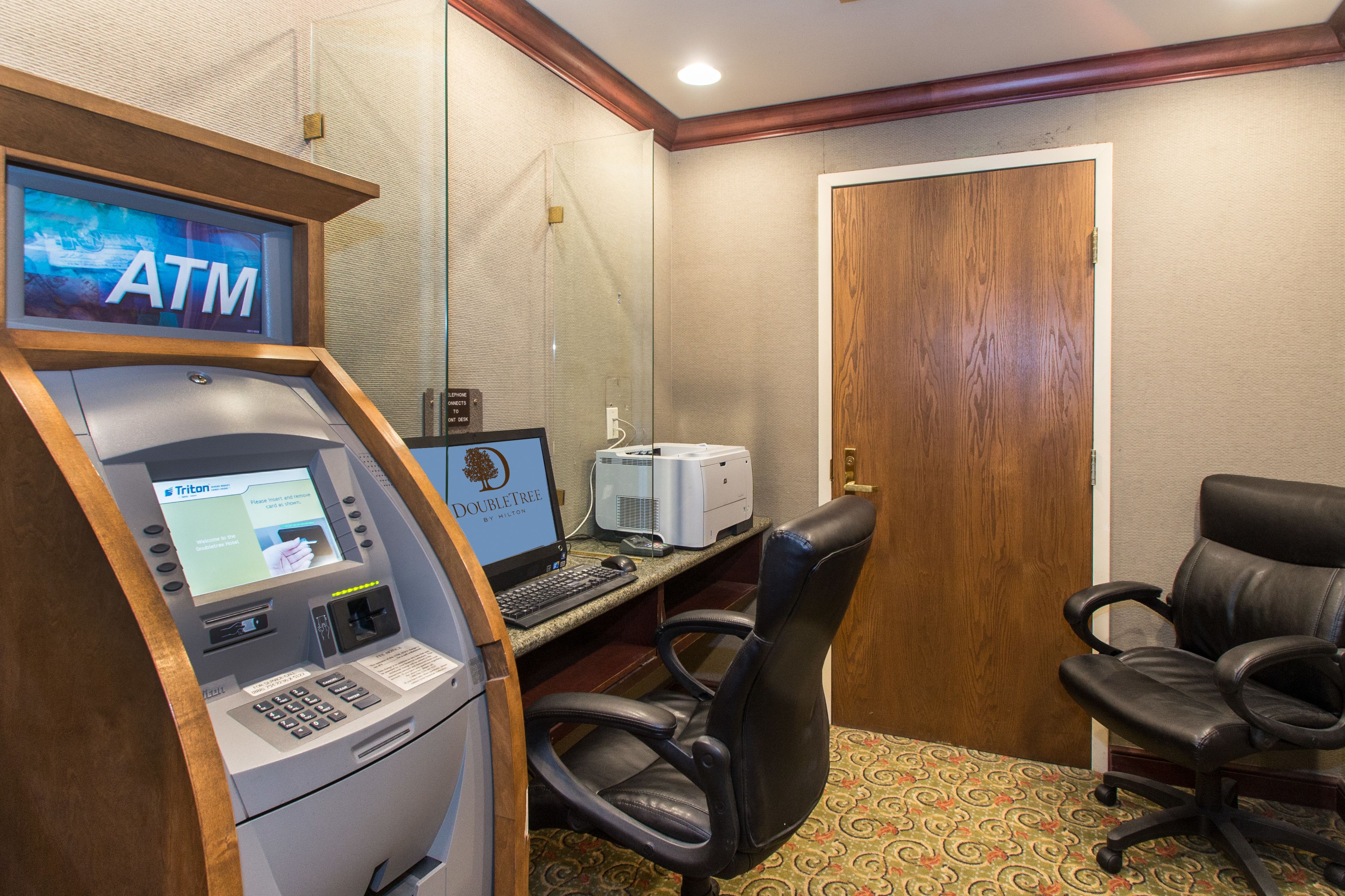 An ATM with a Trilon advertisement, a desktop computer, and two black office chairs in a room with patterned carpet and a wooden door.
