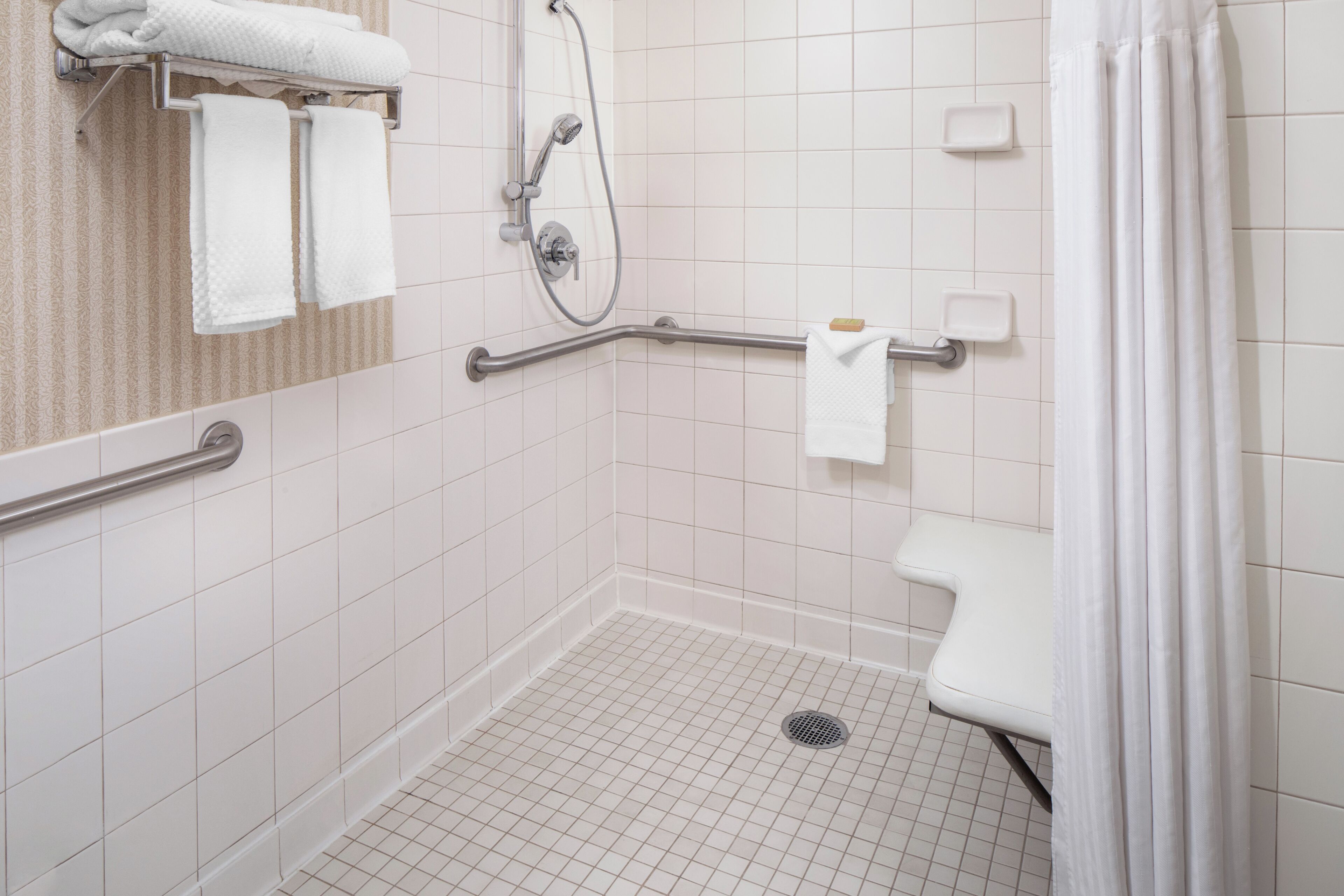 A wheelchair-accessible shower area with grab bars, a white tiled floor, and a white towel rack.