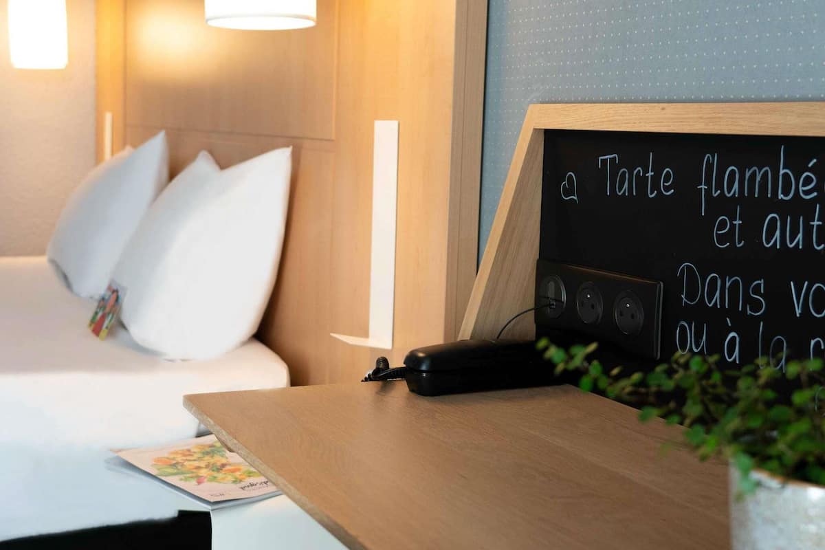 Habitación de hotel con cama, mesita de noche, una pizarra con menú y una lámpara de pared.