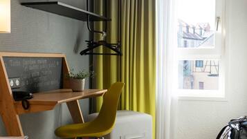 A hotel room with a wooden desk, a yellow chair, a chalkboard with text, a hanging coat rack, and a window with curtains.
