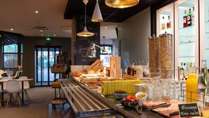 Una cafetería con una barra larga que exhibe pasteles y una variedad de bebidas. Hay mesas y sillas para sentarse, y se ve un menú en una pizarra.