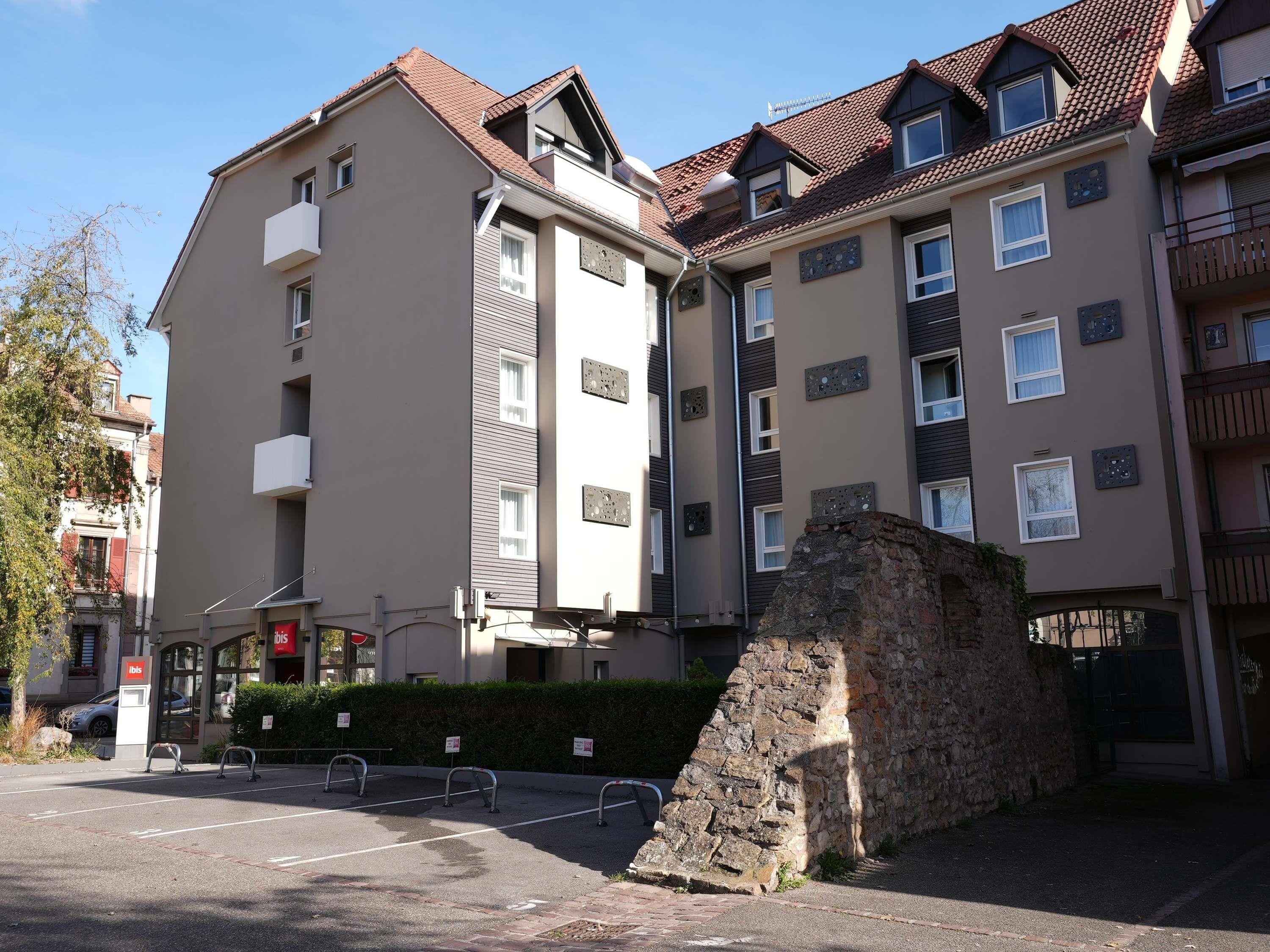 A multi-story residential building with balconies, a stone wall, and a parking area with bollards.