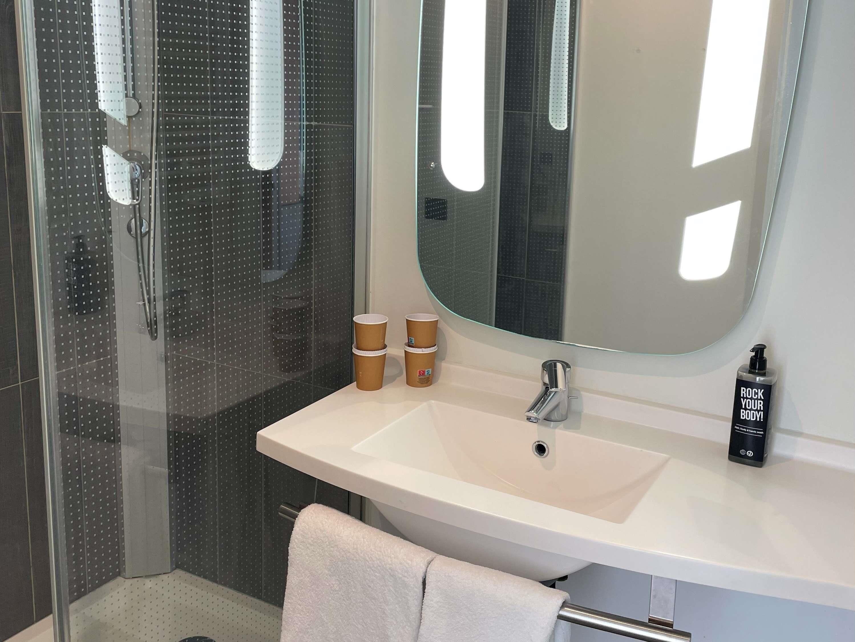 Un baño con cabina de ducha de vidrio, un lavamanos blanco, un espejo grande y una botella de gel de ducha.