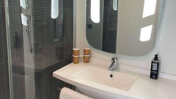 Un baño con cabina de ducha de vidrio, un lavamanos blanco, un espejo grande y una botella de gel de ducha.