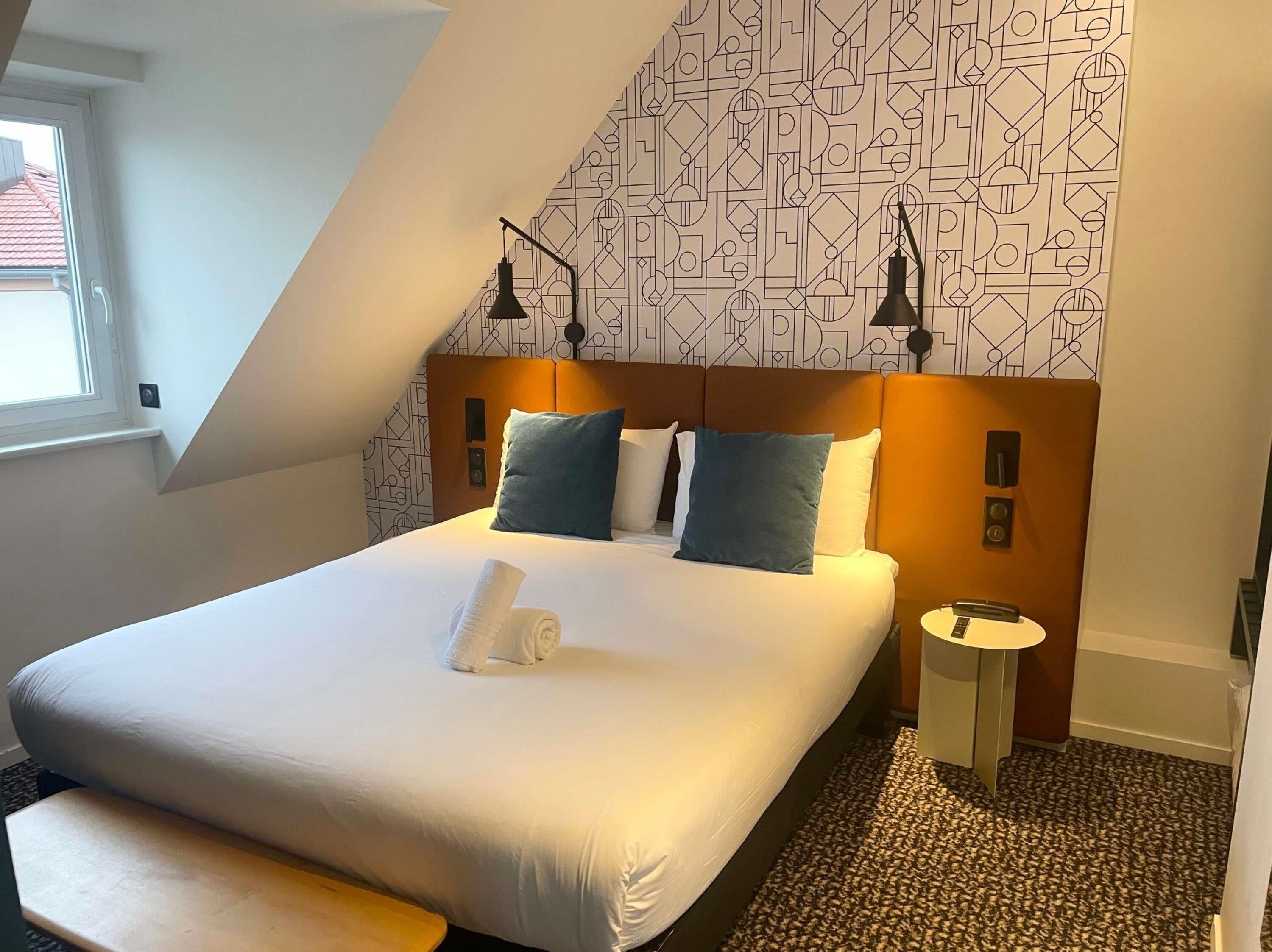 A hotel room with a large bed, a bedside table, a lamp, and a patterned wall.