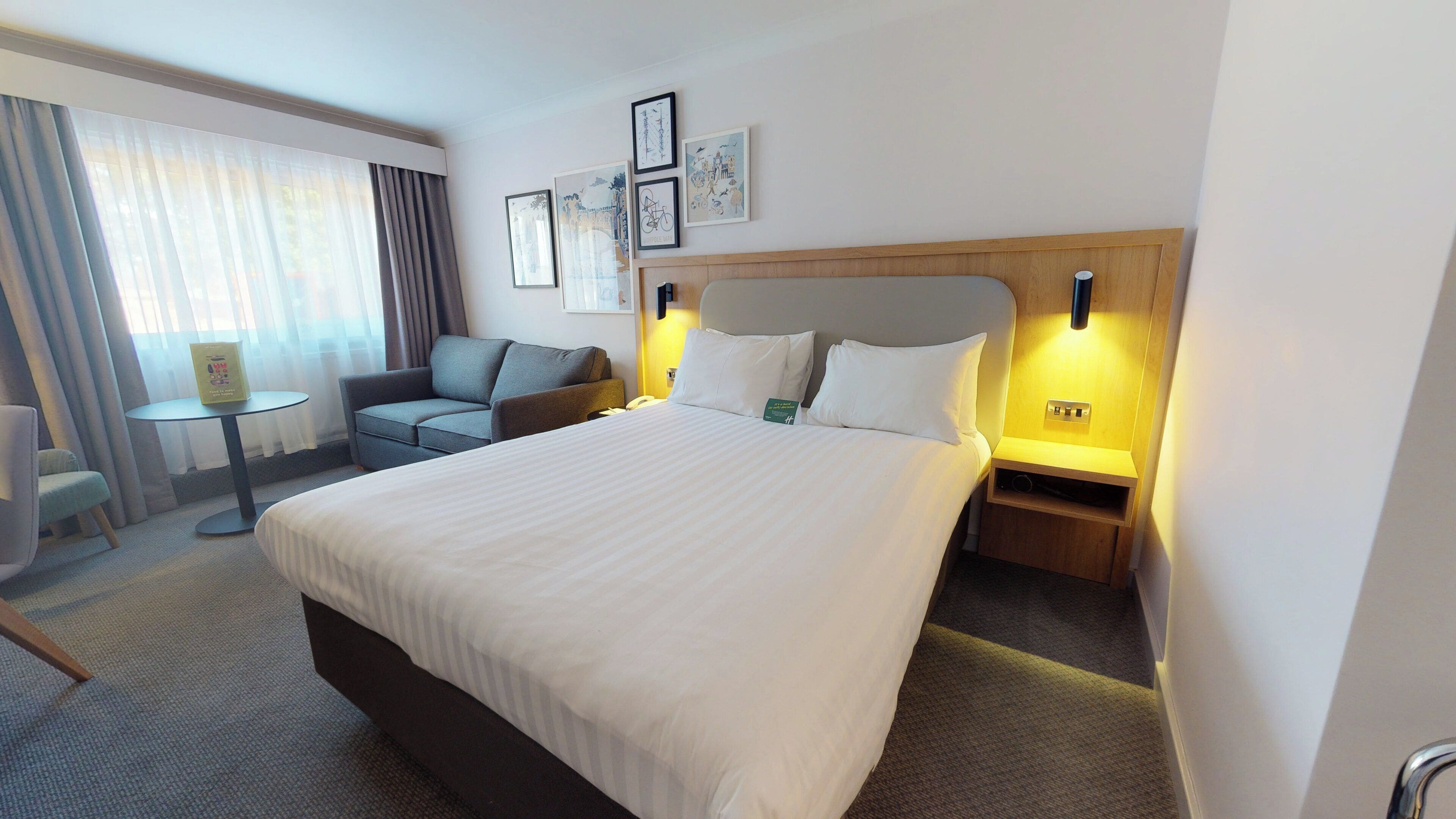 A hotel room with a large bed, a sofa, a small table, and a chair. There are wall-mounted lights and framed pictures on the wall.