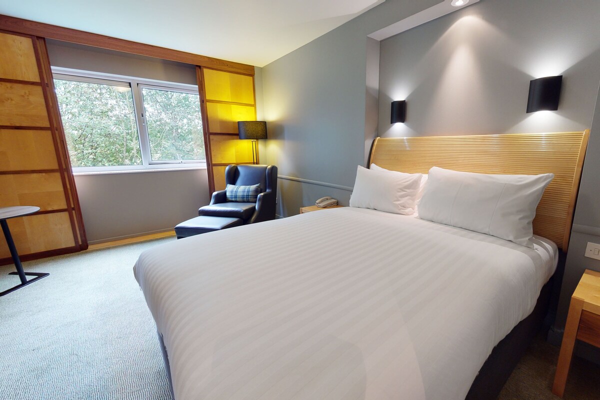 A hotel room with a large bed, a desk, a chair, and a window with a view of trees.