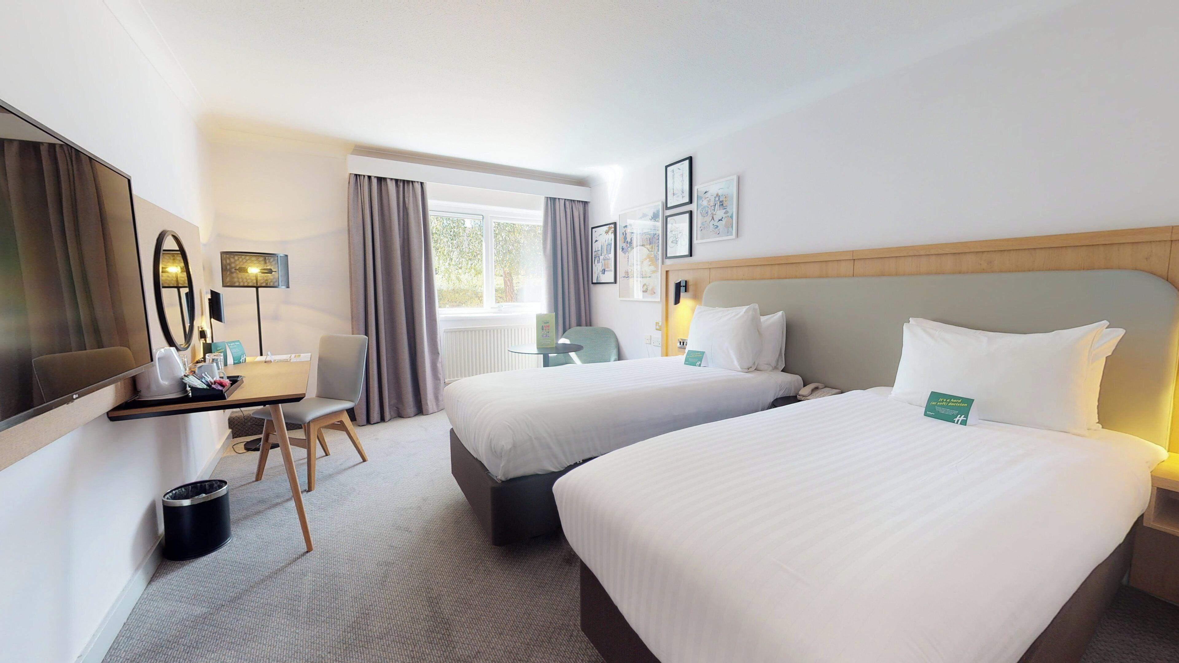 Ein Hotelzimmer mit zwei Betten, einem Schreibtisch mit Stuhl, einem Spiegel und einem fenster mit Vorhängen.