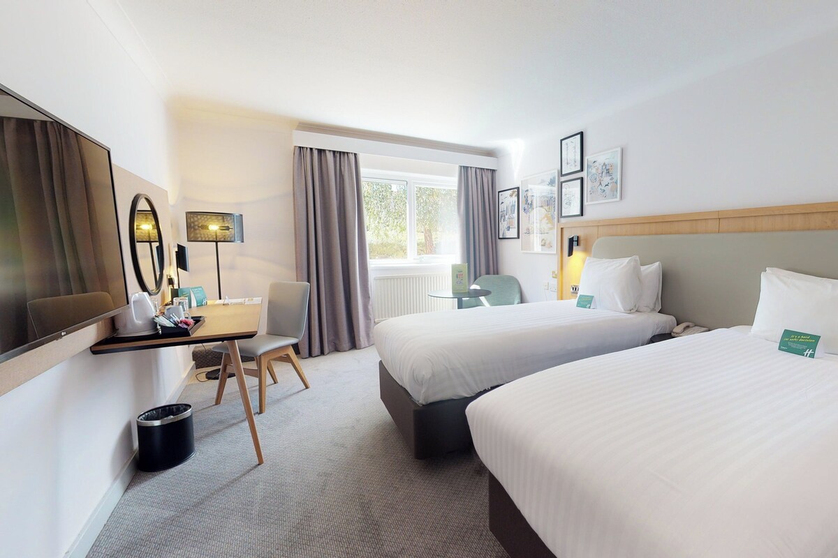 A hotel room with two beds, a desk with a chair, a mirror, and a window with curtains.