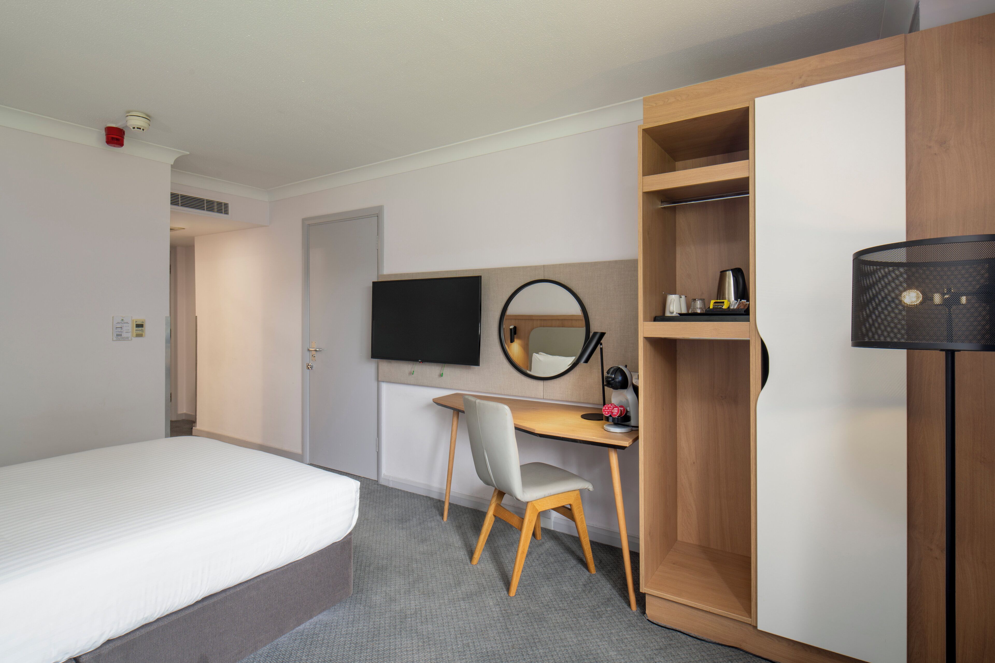 Ein Hotelzimmer mit einem Bett, einem Schreibtisch mit Stuhl, einem Fernseher, einem Kleiderschrank und einem Spiegel.